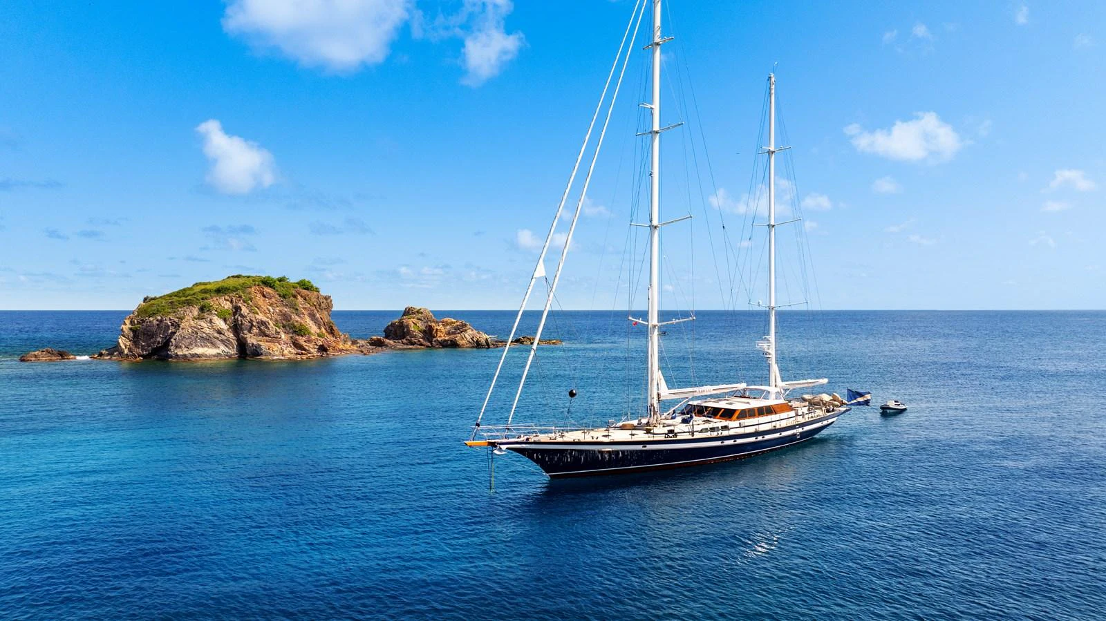 A stunning sailing yacht glides gracefully on calm blue waters near a rocky island. aboard ISLANDIA Yacht for Sale