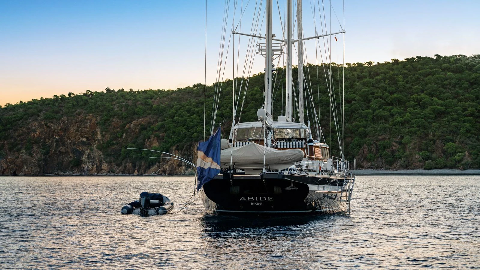A majestic yacht is anchored in serene waters against a backdrop of lush green hills. aboard ISLANDIA Yacht for Sale