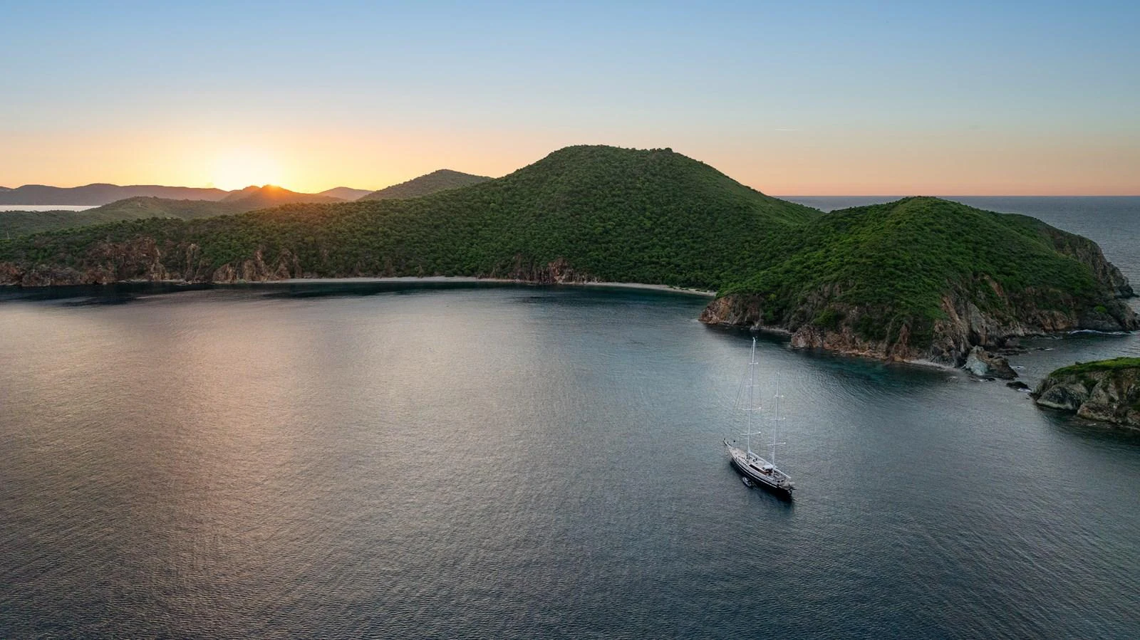 A serene yacht anchored in a tranquil bay at sunset, surrounded by lush green hills. aboard ISLANDIA Yacht for Sale