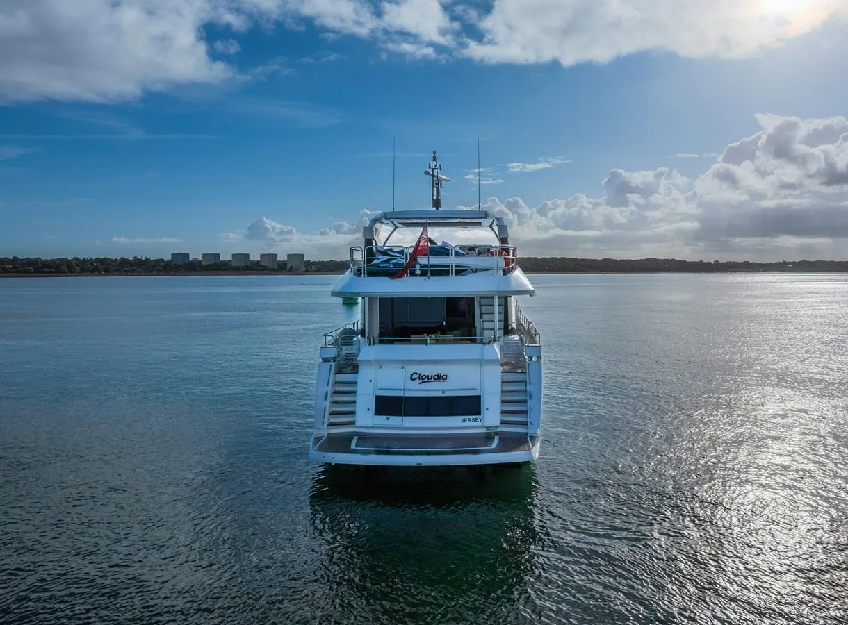 A luxurious yacht is seen from the rear, cruising on calm waters under a partly cloudy sky. aboard CLOUDIA Yacht for Sale
