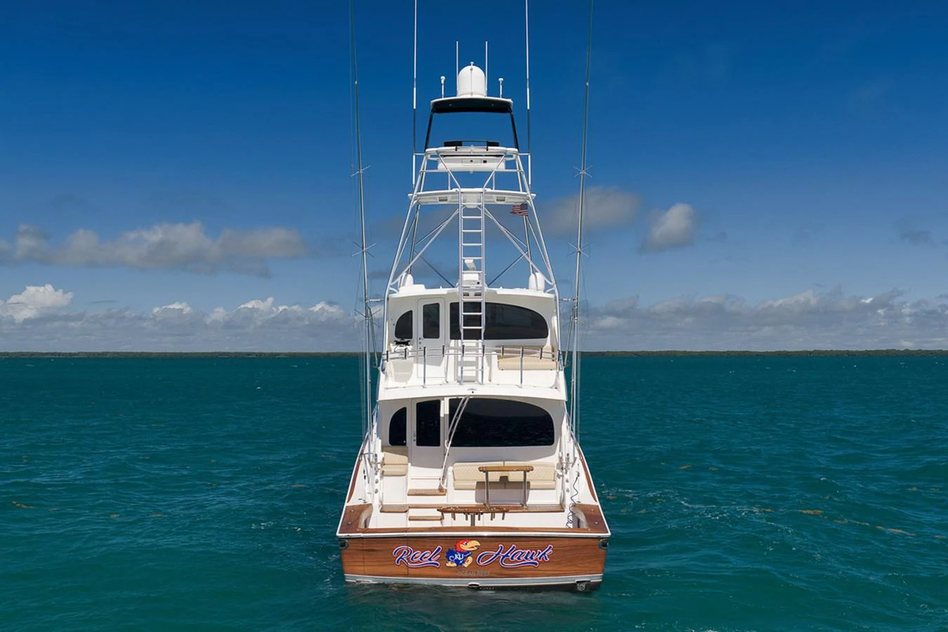 The yacht is seen from the rear, showcasing its multi-level structure and open deck against a serene ocean backdrop. aboard REEL HAWK Yacht for Sale