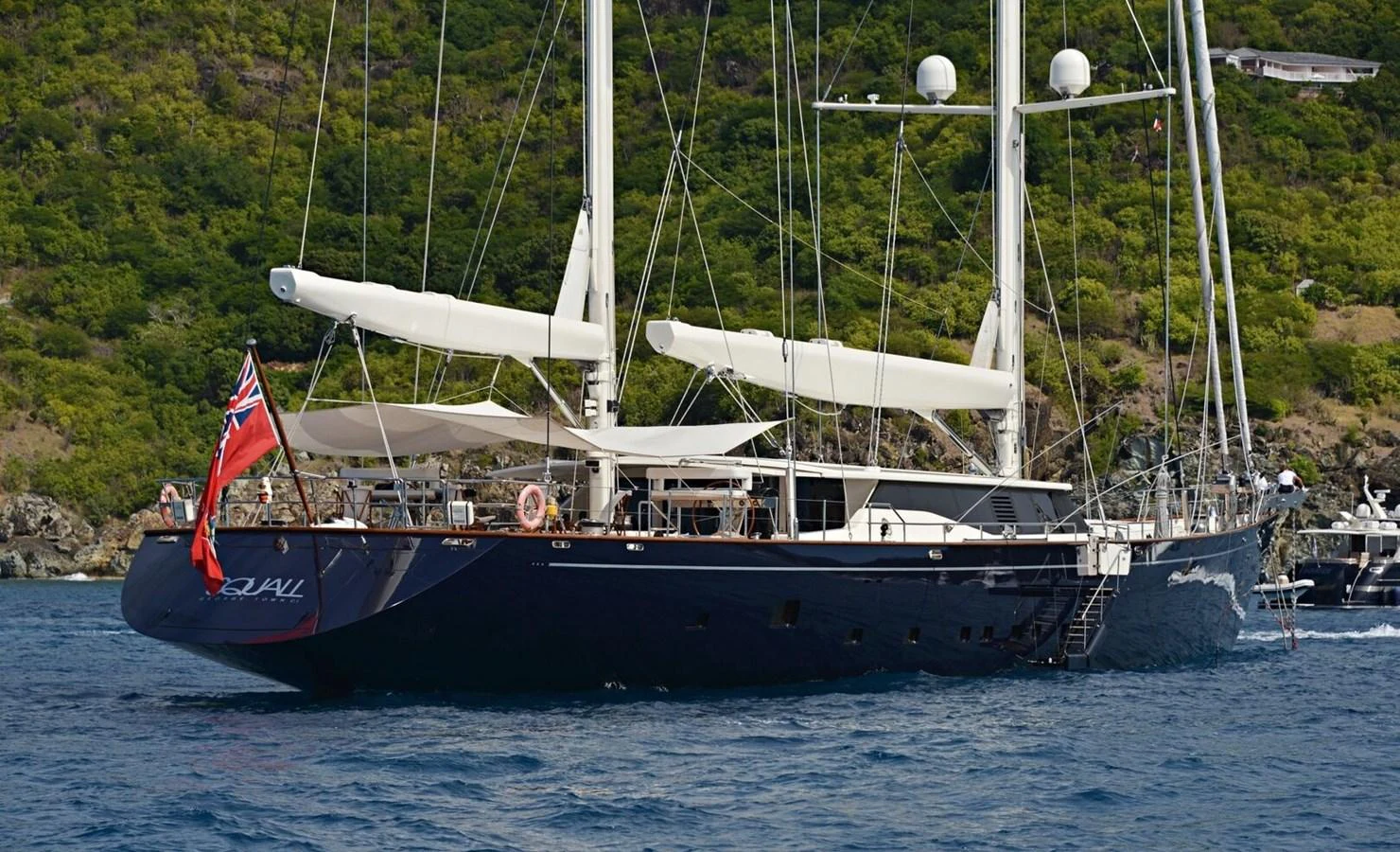 A sleek sailing yacht glides gracefully on the water with lush green hills in the background. aboard SQUALL Yacht for Sale