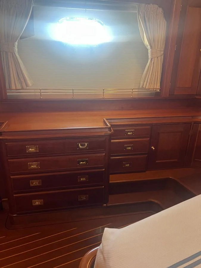 The image showcases a classic wooden interior with a chest of drawers and a window with curtains. aboard KNICKERBOCKER Yacht for Sale