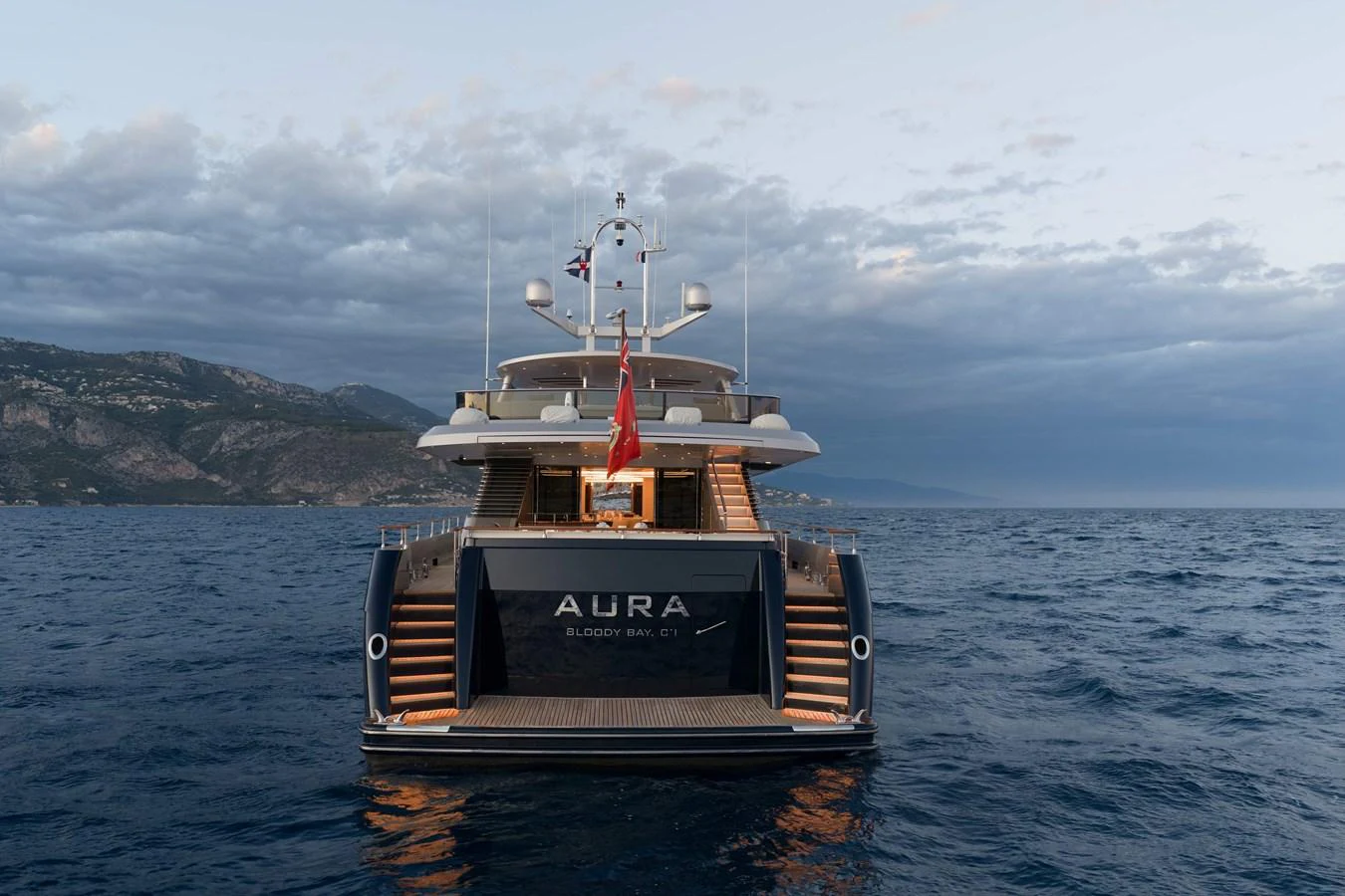 The yacht is captured from the stern, showcasing its elegant design and spacious aft deck against a scenic coastal backdrop. aboard AURA Yacht for Sale