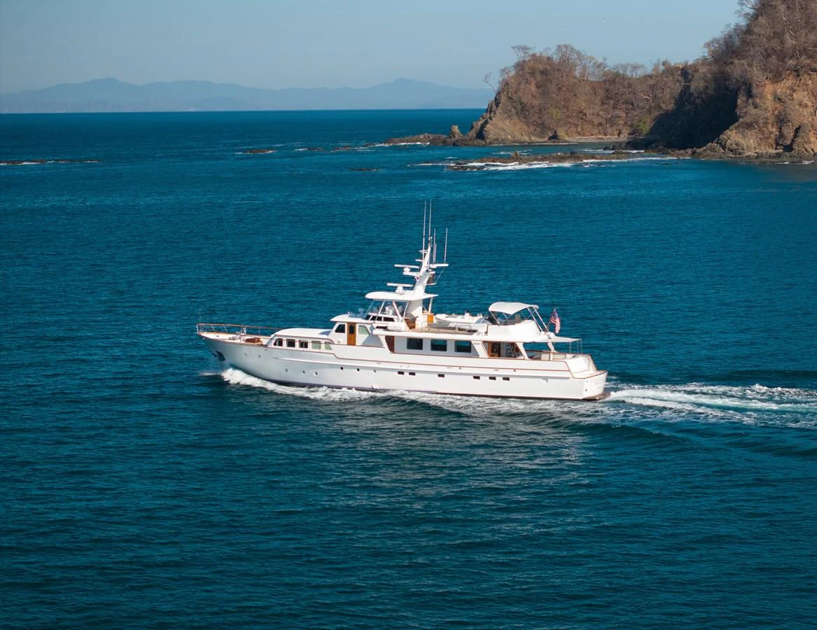A classic yacht cruises gracefully through clear blue waters near a rocky coastline. aboard PASSAGE Yacht for Sale