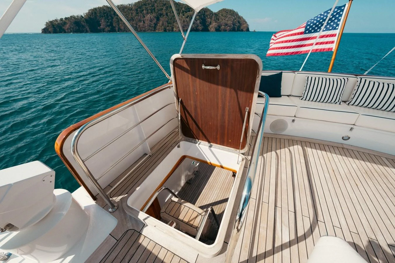 The aft deck features a hatch leading below deck, with a scenic view of the ocean and an American flag waving in the background. aboard PASSAGE Yacht for Sale