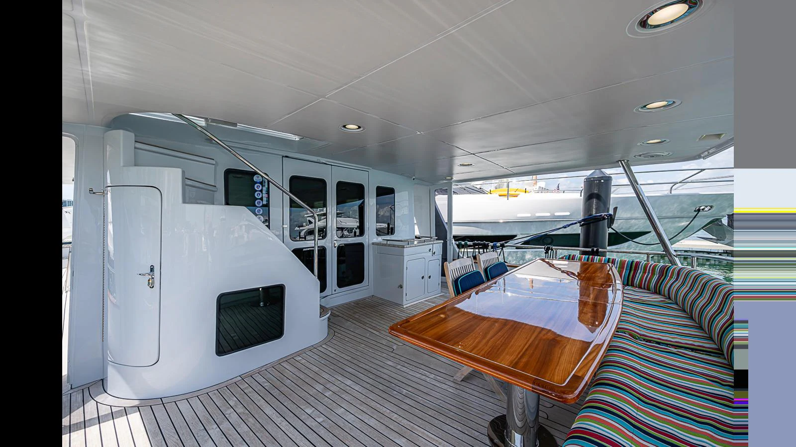 The aft deck features a spacious seating area with a polished wooden table and a curved bench with colorful cushions. aboard TURANGALILA Yacht for Sale