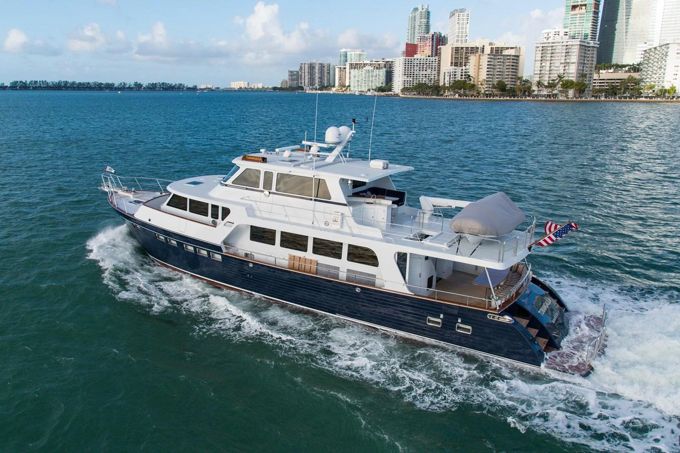 A classic motor yacht cruises through vibrant blue waters with a city skyline in the background. aboard TURANGALILA Yacht for Sale