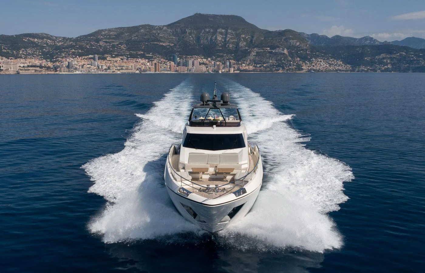 A sleek yacht cruises through calm waters with a picturesque coastal city and mountains in the background. aboard ALEGRIA II Yacht for Sale