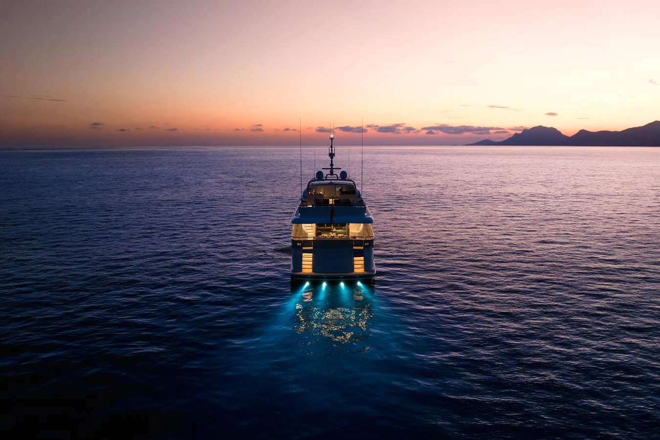 A yacht is illuminated by underwater lights as it sails into the sunset. aboard ORIZZONTE Yacht for Sale