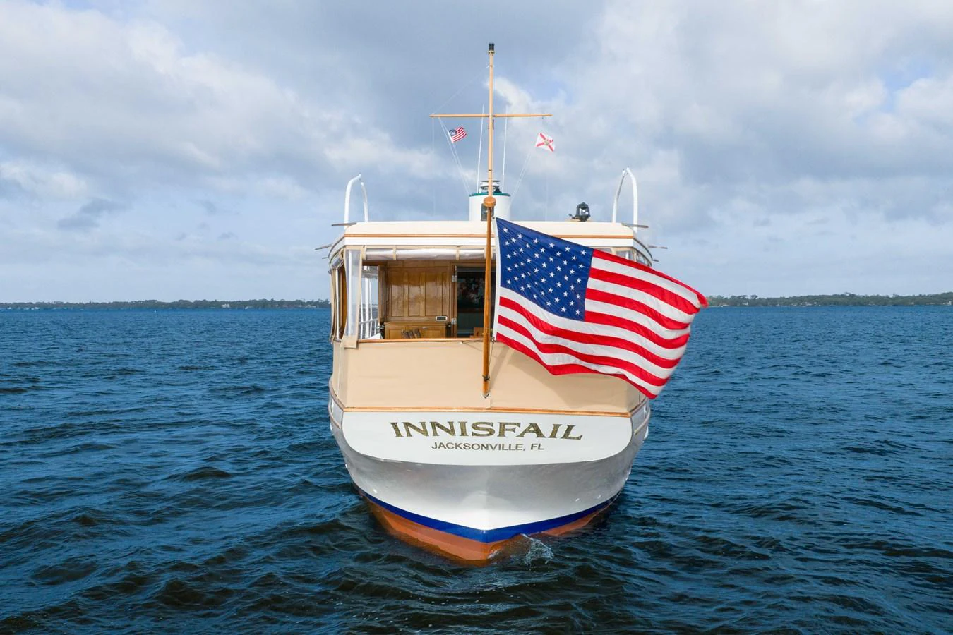 A classic yacht with an American flag cruises on calm waters under a cloudy sky. aboard INNISFAIL Yacht for Sale