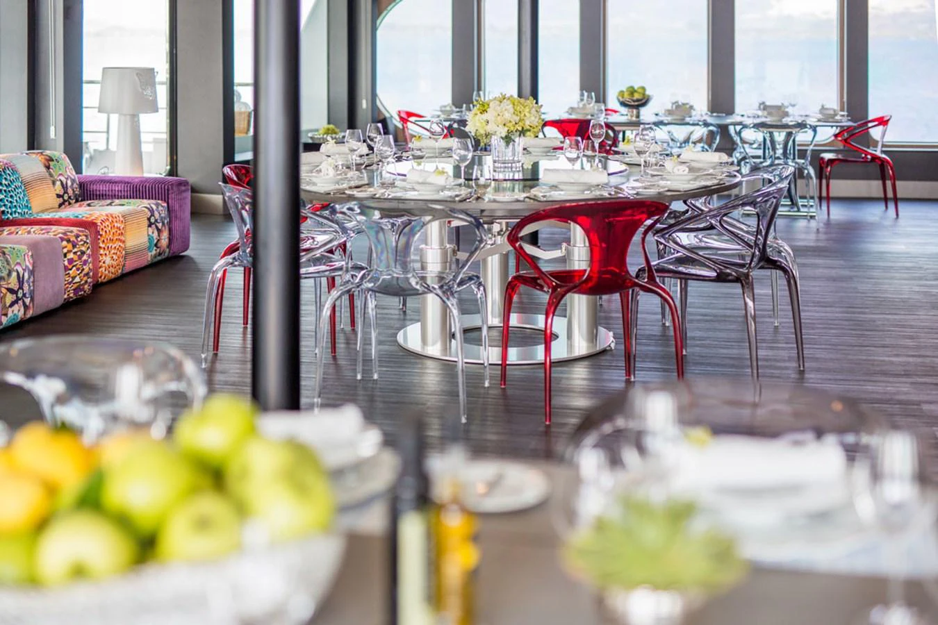 The yacht's dining area features modern furnishings with a mix of transparent and red chairs around a large table, complemented by a vibrant sofa and panoramic windows. aboard SALUZI Yacht for Sale