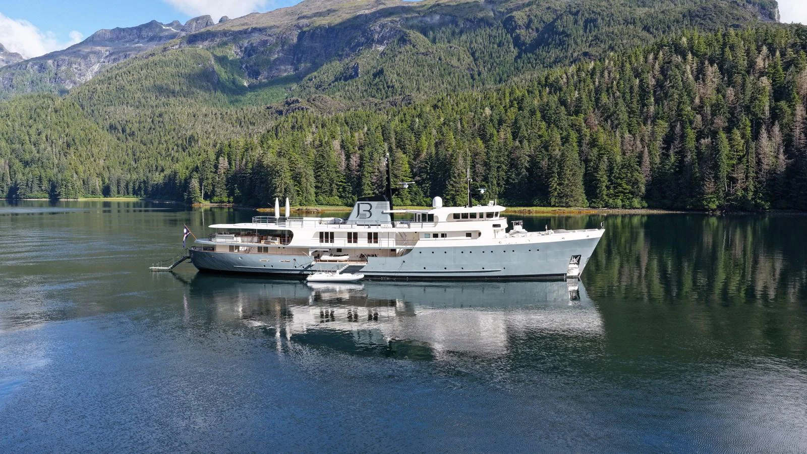 A luxurious yacht is anchored in a serene, forested bay with mountains in the background. aboard JUST B Yacht for Sale