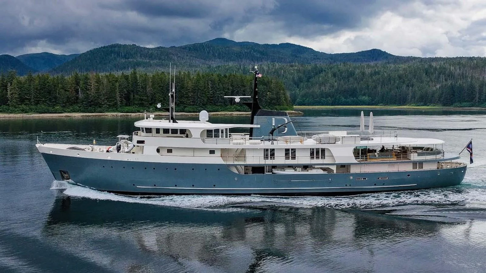 A majestic yacht cruises through calm waters with a scenic forested backdrop. aboard JUST B Yacht for Sale