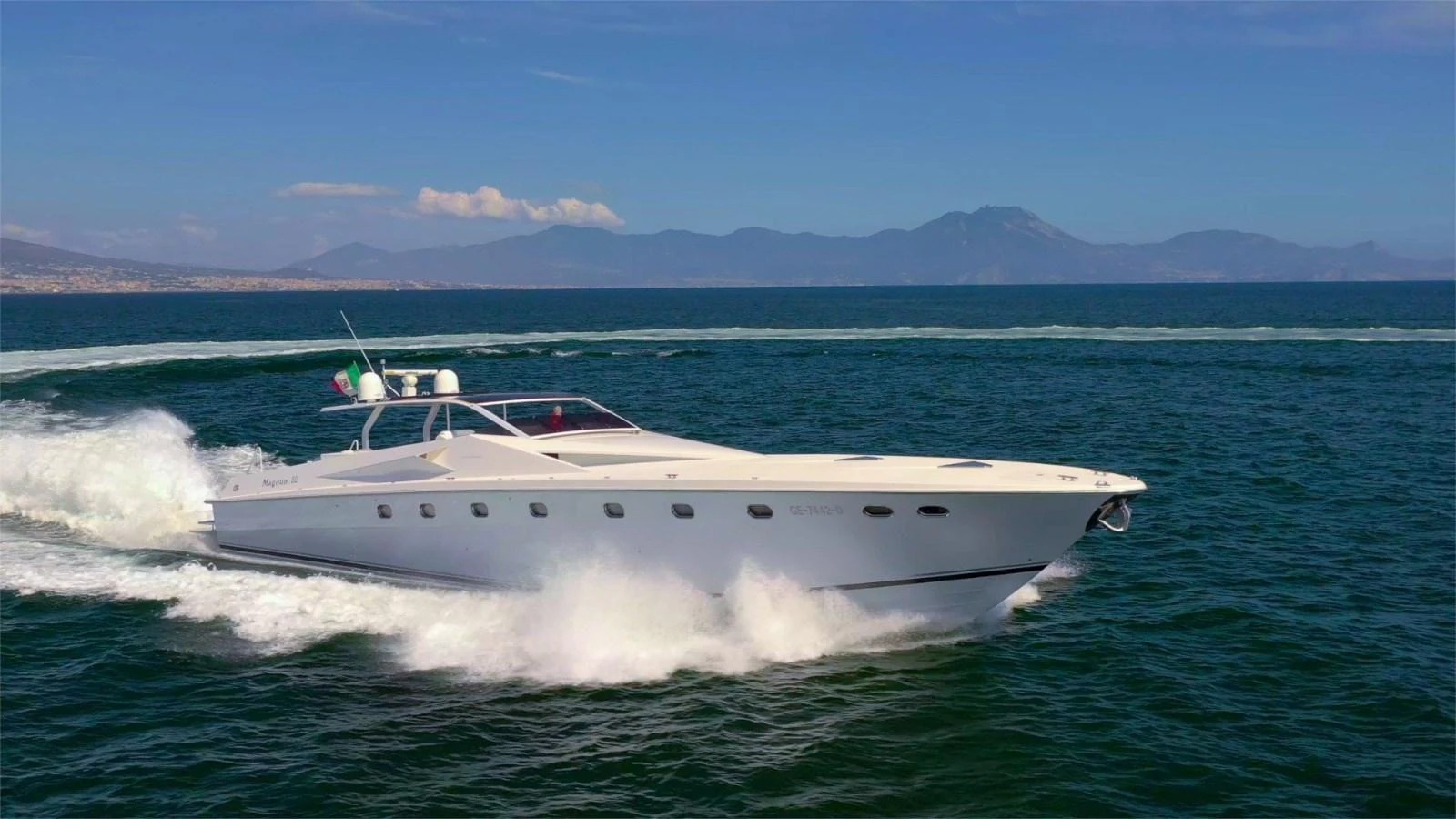 A sleek, white yacht cruises swiftly across the open sea with a mountainous backdrop. aboard WHITE & SILVER Yacht for Sale