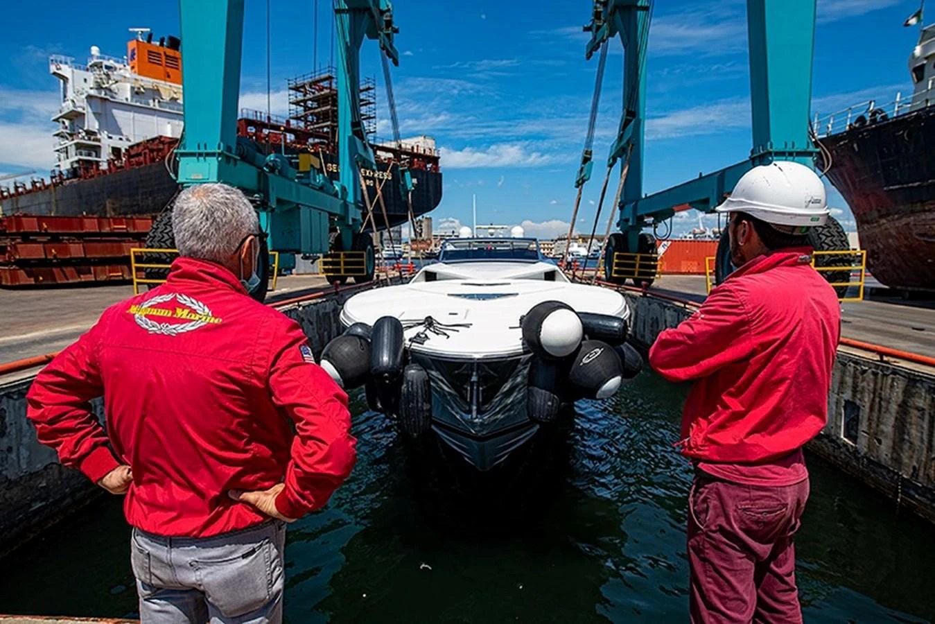A yacht is being lowered into the water at a shipyard, overseen by two workers in red jackets. aboard WHITE & SILVER Yacht for Sale