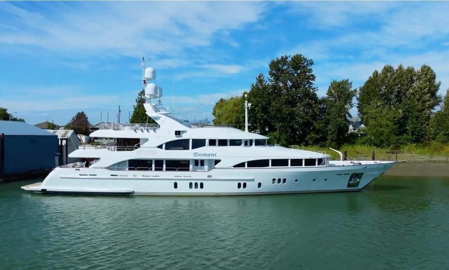 A sleek and elegant yacht is docked in calm waters, showcasing its luxurious design against a backdrop of trees and blue sky. aboard DOMANI Yacht for Sale