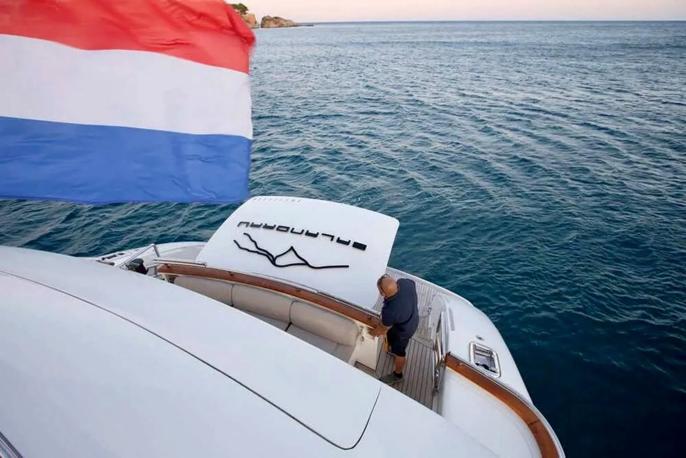 A view of the yacht's aft deck with a person standing near the seating area and a flag waving in the breeze. aboard BALANDRAU Yacht for Sale