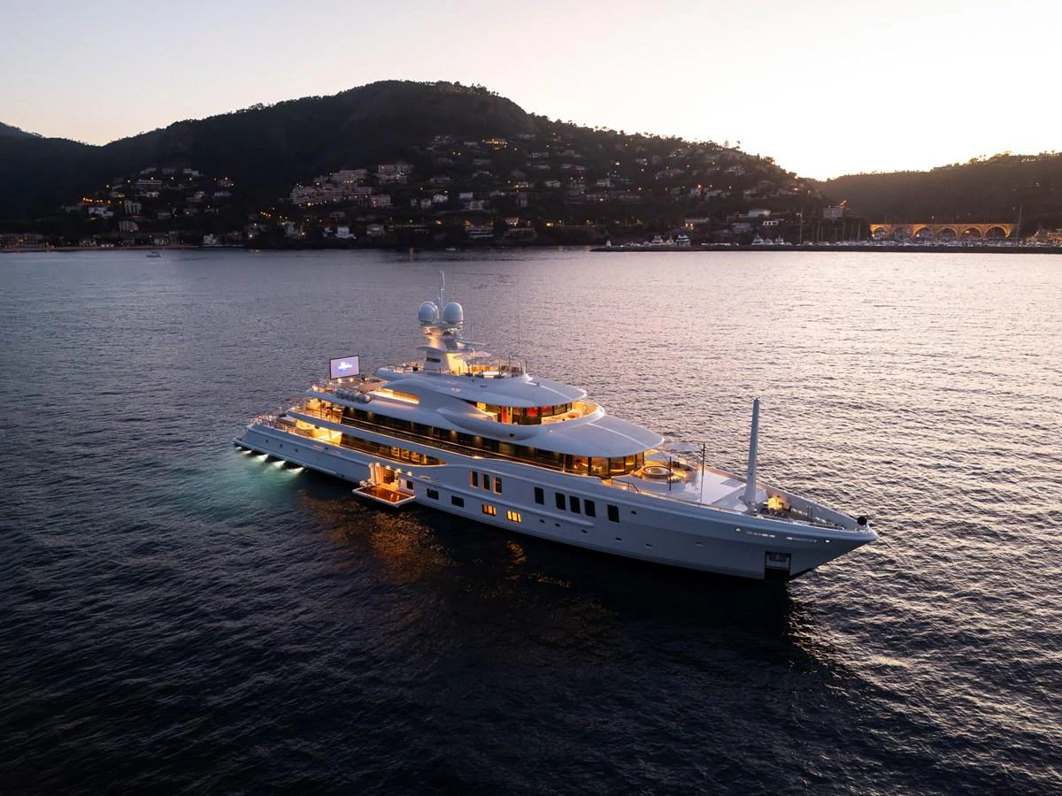 A luxurious yacht is elegantly illuminated against a serene coastal backdrop at dusk. aboard SYNTHESIS Yacht for Sale