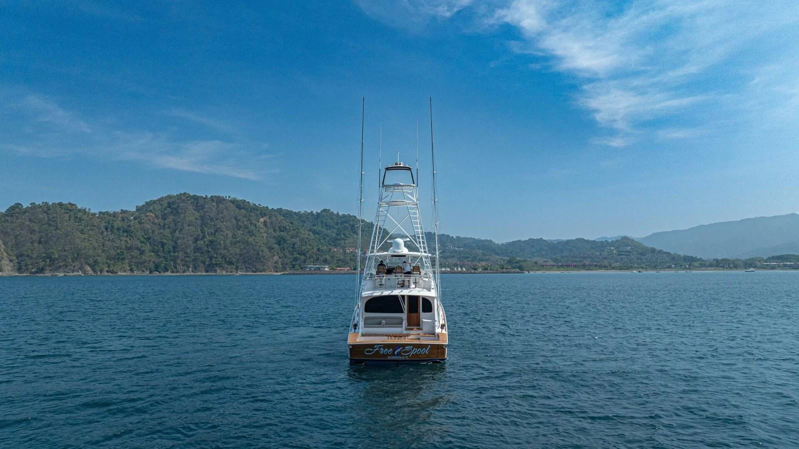 A yacht is pictured from the rear, cruising on calm waters with a scenic mountainous backdrop. aboard FREE SPOOL Yacht for Sale