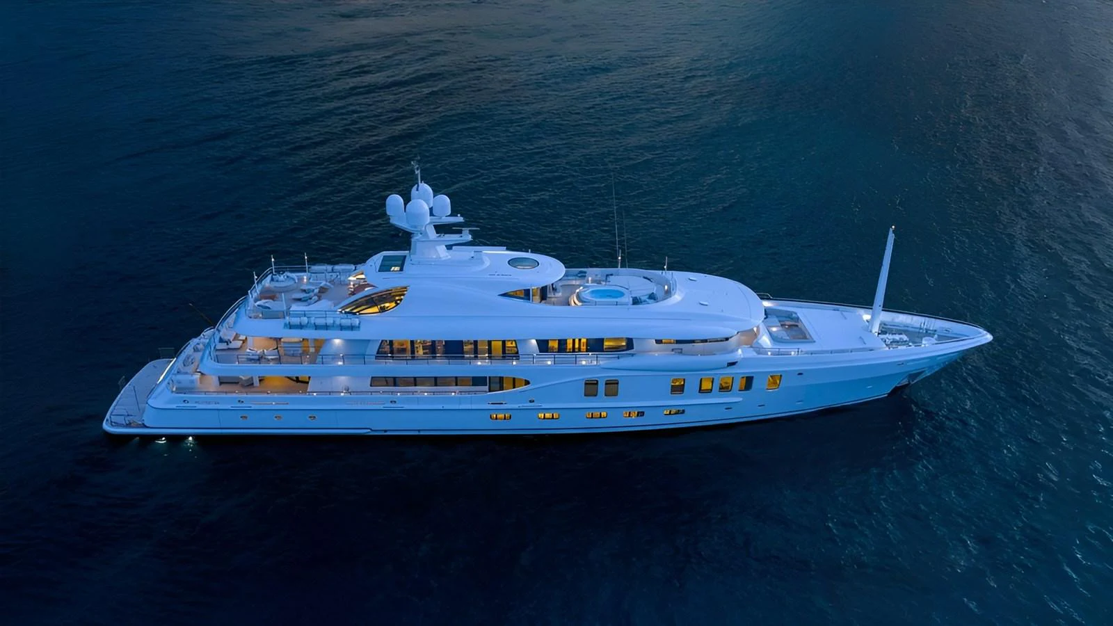 A stunning superyacht glides gracefully on calm waters at dusk, illuminated by soft, warm lights. aboard NENINKA Yacht for Sale
