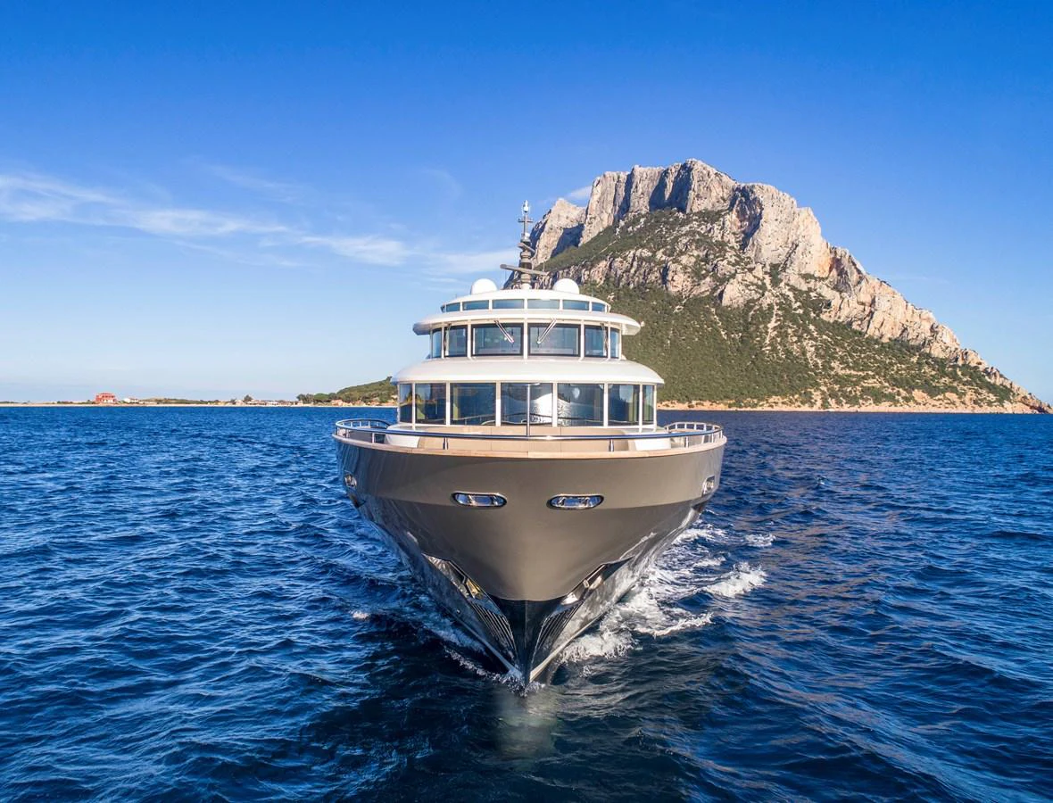 A luxurious yacht cruises through clear blue waters with a stunning mountainous backdrop. aboard MAXIMA Yacht for Sale