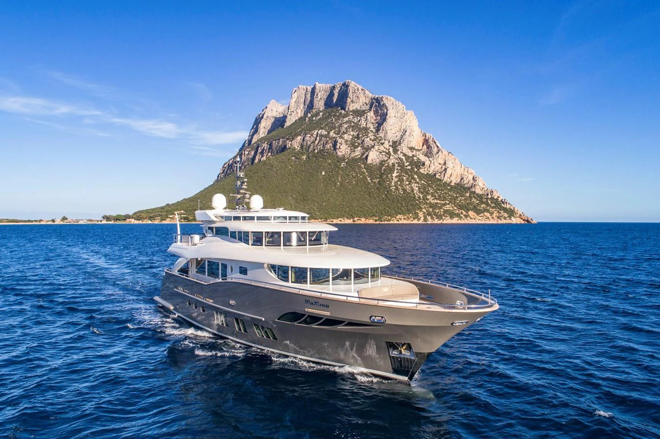 A luxurious yacht cruises through pristine blue waters with a stunning mountainous backdrop. aboard MAXIMA Yacht for Sale