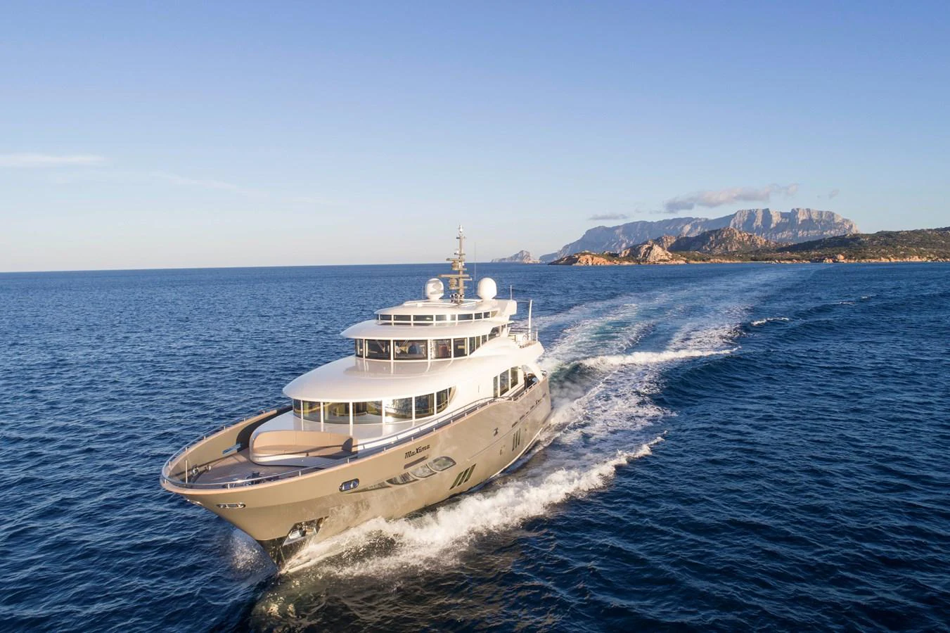 A luxurious yacht cruises through clear blue waters with a scenic coastline in the background. aboard MAXIMA Yacht for Sale