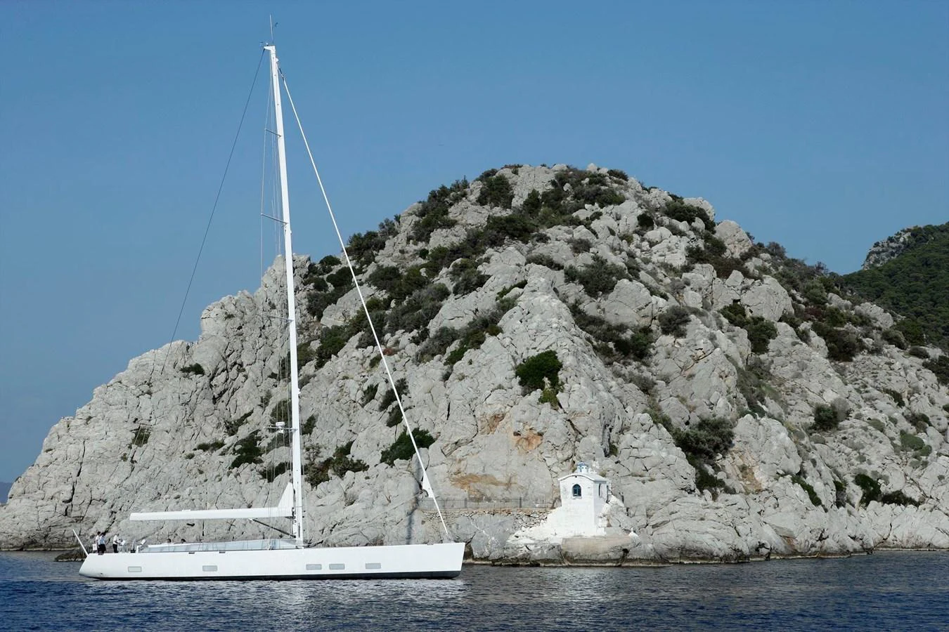 A sleek yacht sails near a rocky coastline under a clear blue sky. aboard S/Y SENTINEL Yacht for Sale