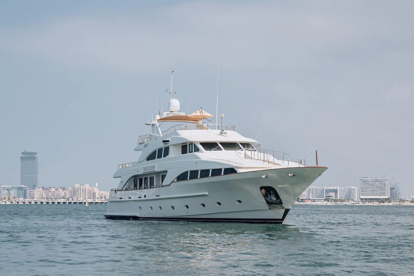 A luxurious yacht glides gracefully on calm waters with a cityscape in the background. aboard GALLUS Yacht for Sale