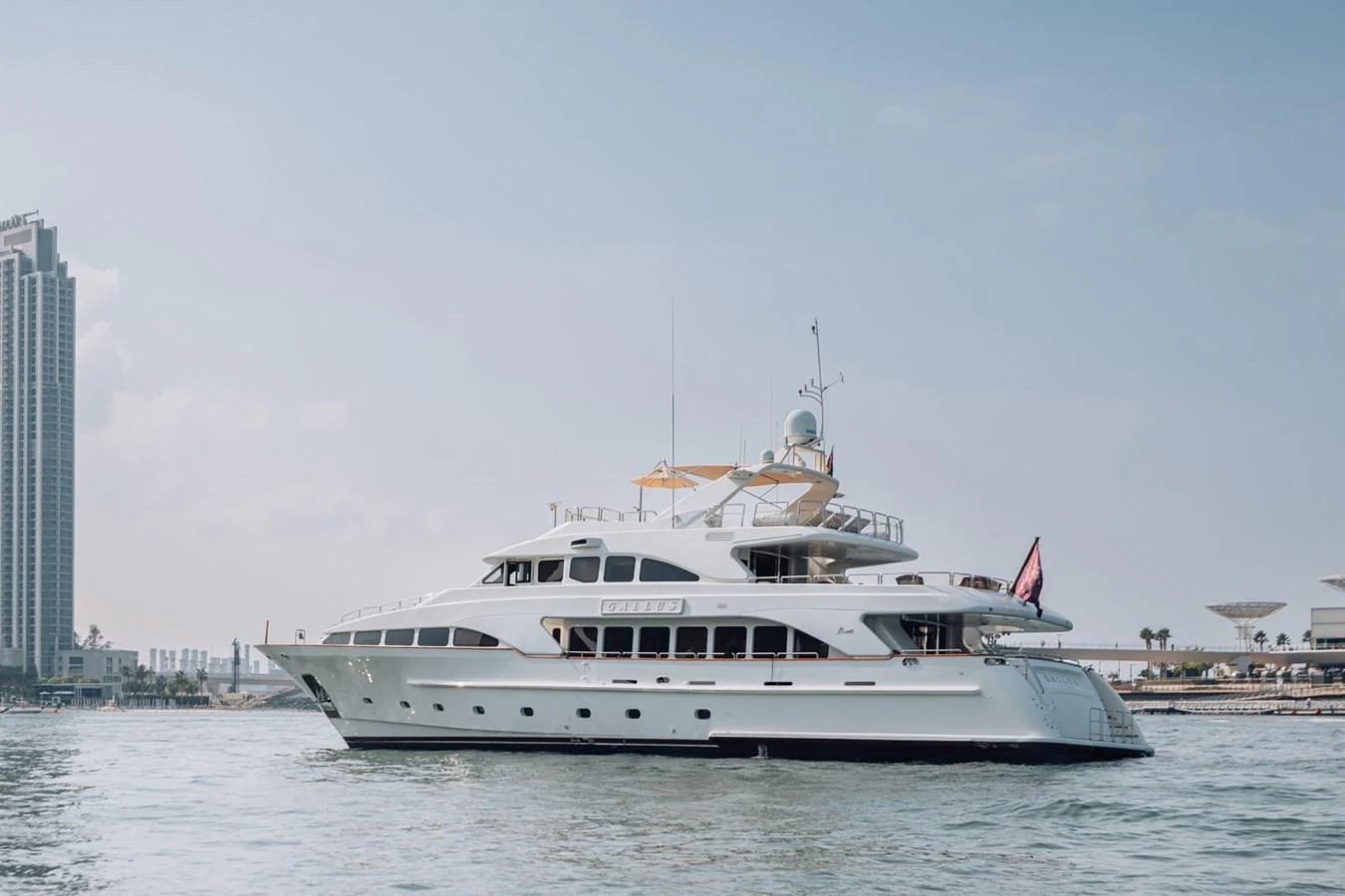 A luxurious yacht glides gracefully on calm waters with a cityscape in the background. aboard GALLUS Yacht for Sale