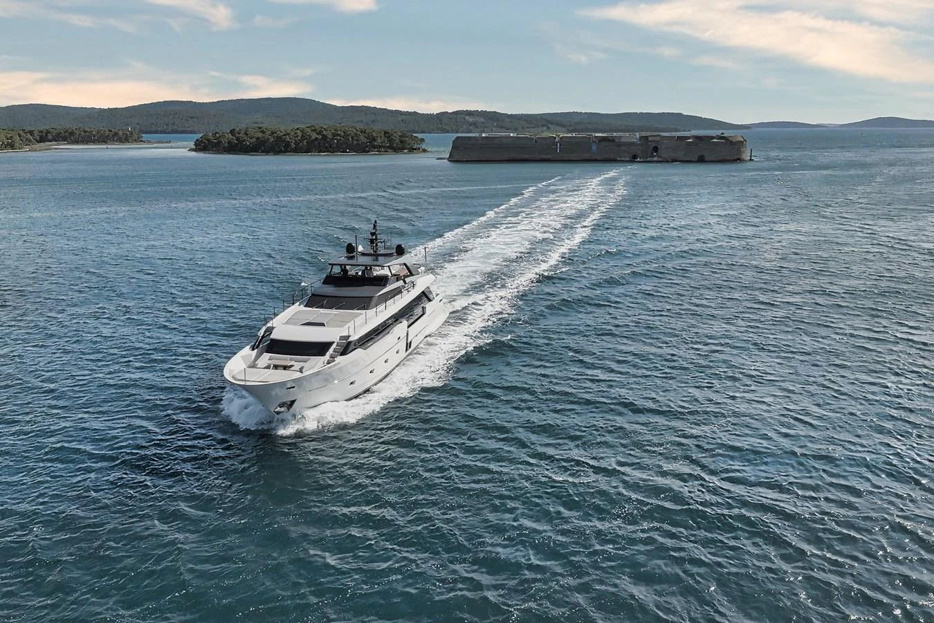 A sleek yacht cruises through calm waters with a scenic backdrop of islands and a fortress. aboard DESTINO Yacht for Sale