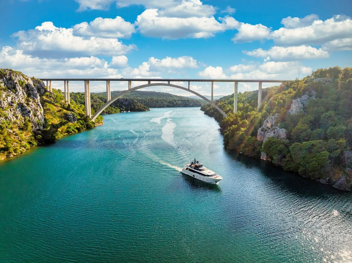 A luxurious yacht cruises through a picturesque river under a grand bridge, surrounded by lush greenery and clear skies. aboard DESTINO Yacht for Sale
