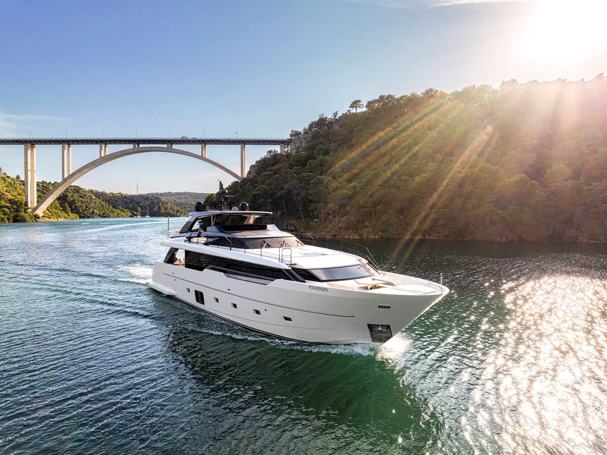 A sleek yacht cruises through a scenic waterway with a bridge in the background, bathed in sunlight. aboard DESTINO Yacht for Sale