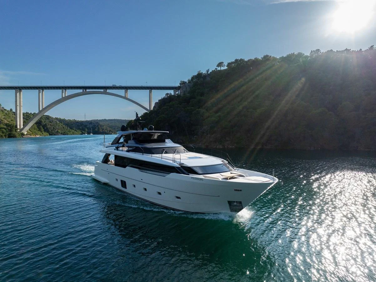 A sleek yacht cruises through a scenic waterway under a bridge, bathed in sunlight. aboard DESTINO Yacht for Sale