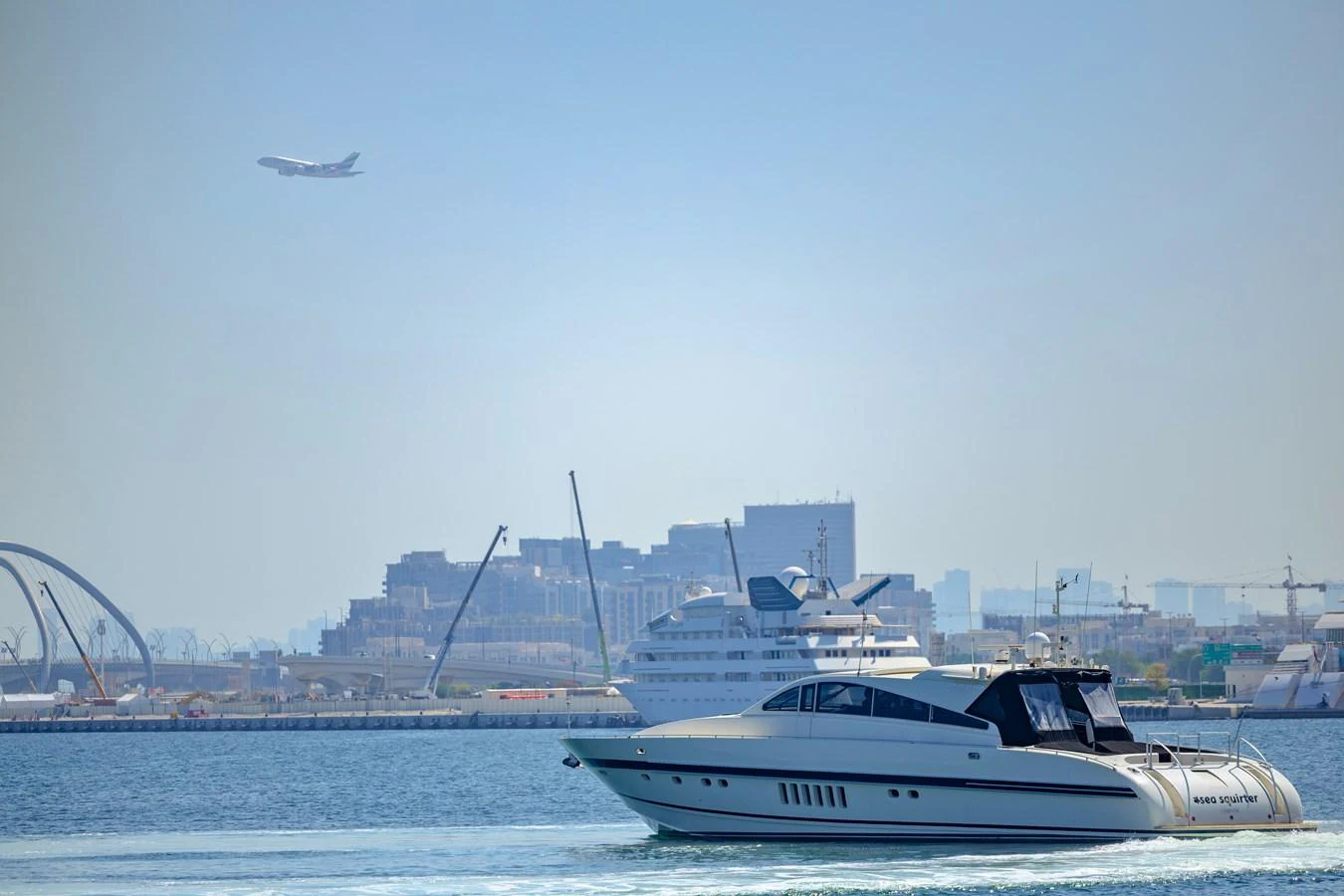 A sleek yacht cruises through calm waters with a cityscape and another yacht in the background. aboard SEA SQUIRTER Yacht for Sale