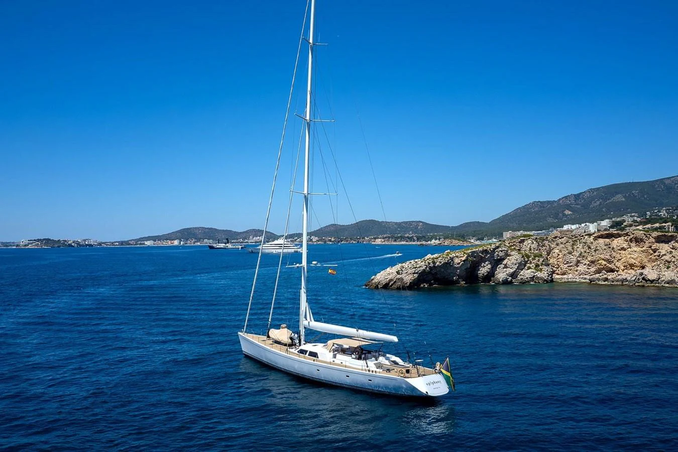 A sleek sailing yacht glides gracefully on calm blue waters near a rocky coastline. aboard EPIPHANY Yacht for Sale