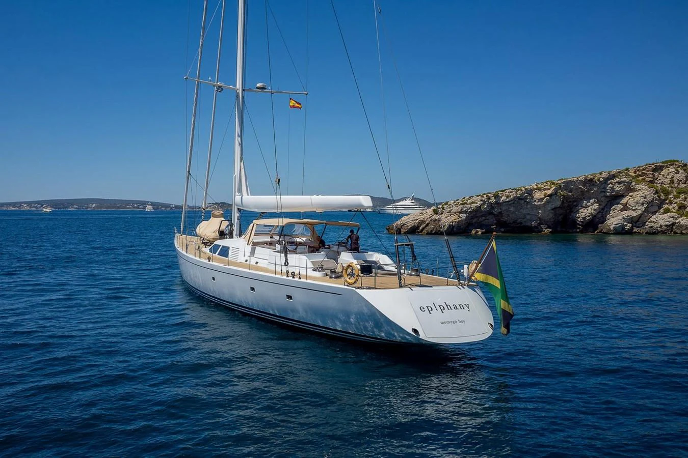 A sleek sailing yacht glides gracefully on calm blue waters near a rocky coastline. aboard EPIPHANY Yacht for Sale