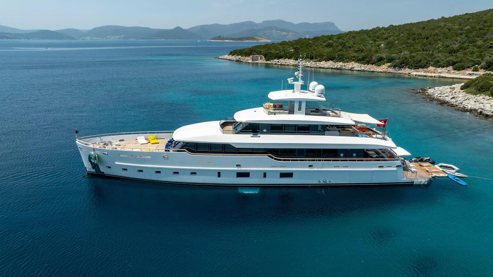 A sleek and modern yacht is anchored in a serene, turquoise bay with lush green hills in the background. aboard K Yacht for Sale