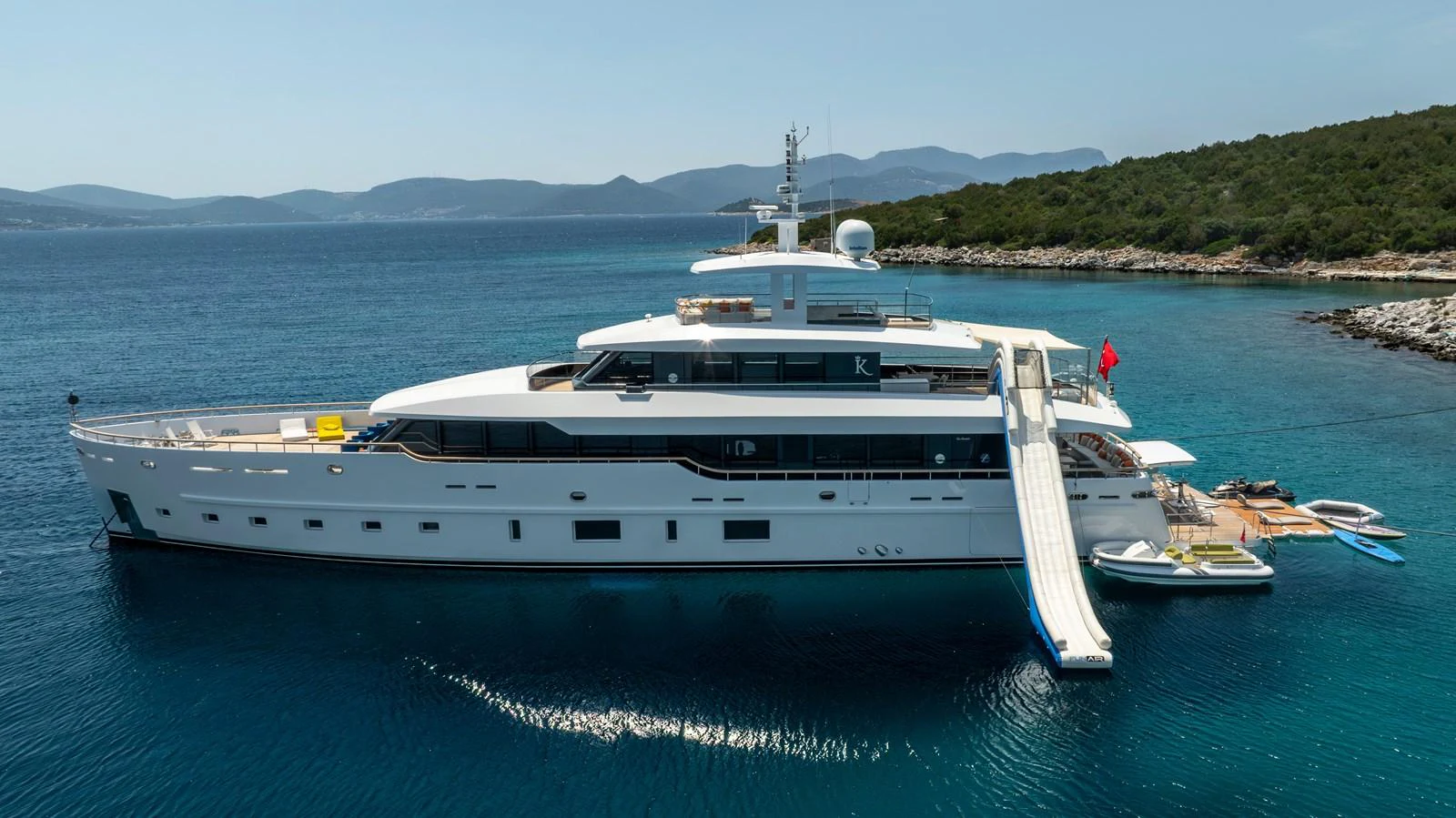 A sleek and modern yacht anchored in a serene coastal area with a waterslide extending from the deck. aboard K Yacht for Sale