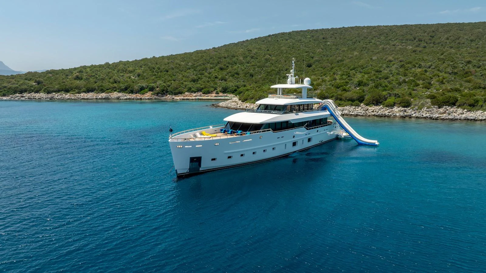 A luxurious yacht anchored in a serene bay with a water slide extending from the upper deck. aboard K Yacht for Sale