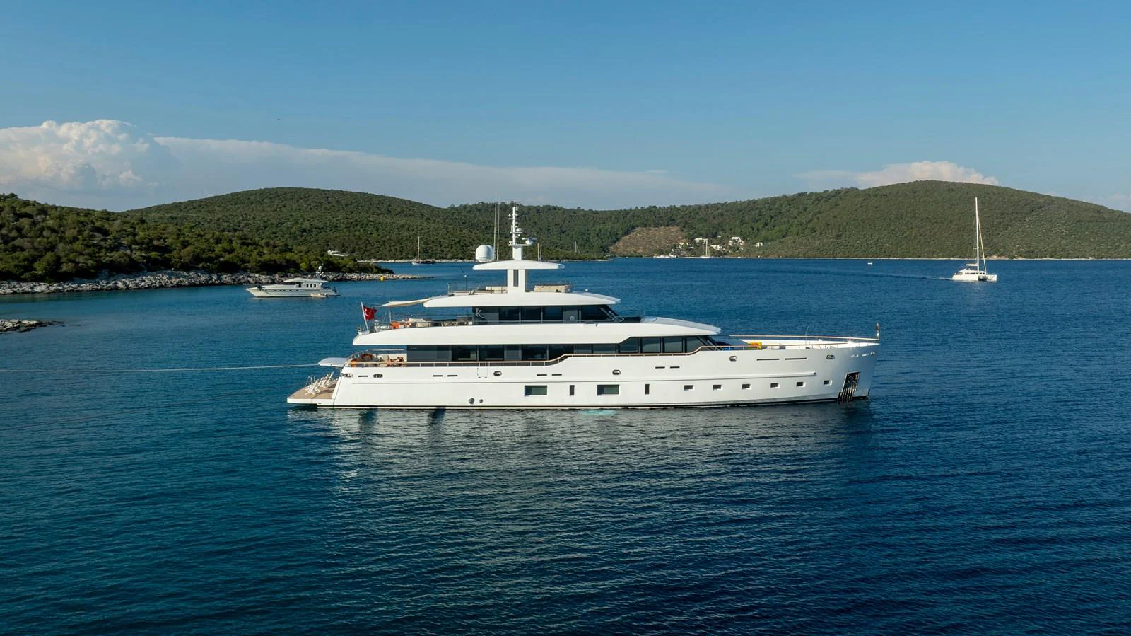 A sleek and modern yacht is anchored in a serene bay with clear blue waters and lush green hills in the background. aboard K Yacht for Sale