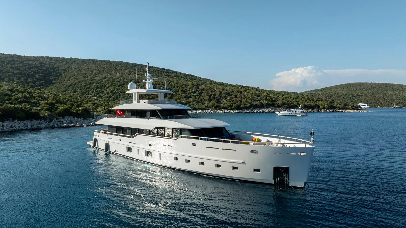 A sleek, modern yacht glides through serene blue waters with lush green hills in the background. aboard K Yacht for Sale