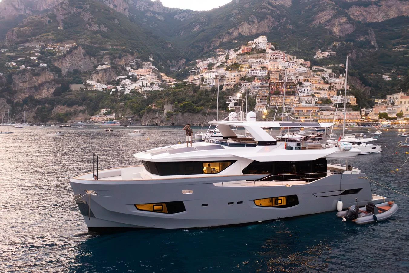 A sleek yacht is anchored in a picturesque bay with a charming hillside village in the background. aboard KAMOKA Yacht for Sale