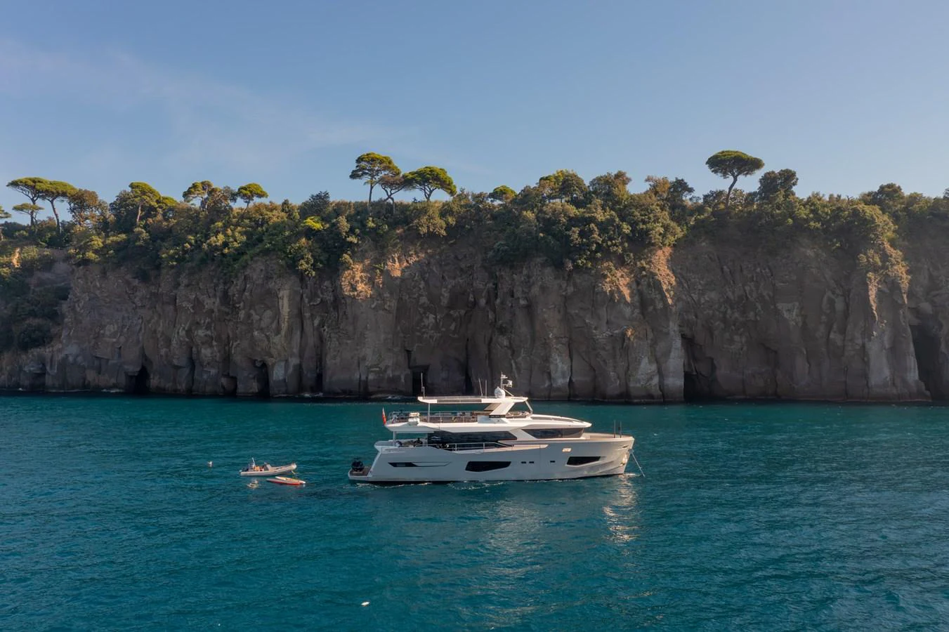 A luxurious yacht is anchored near a scenic rocky coastline with lush greenery. aboard KAMOKA Yacht for Sale