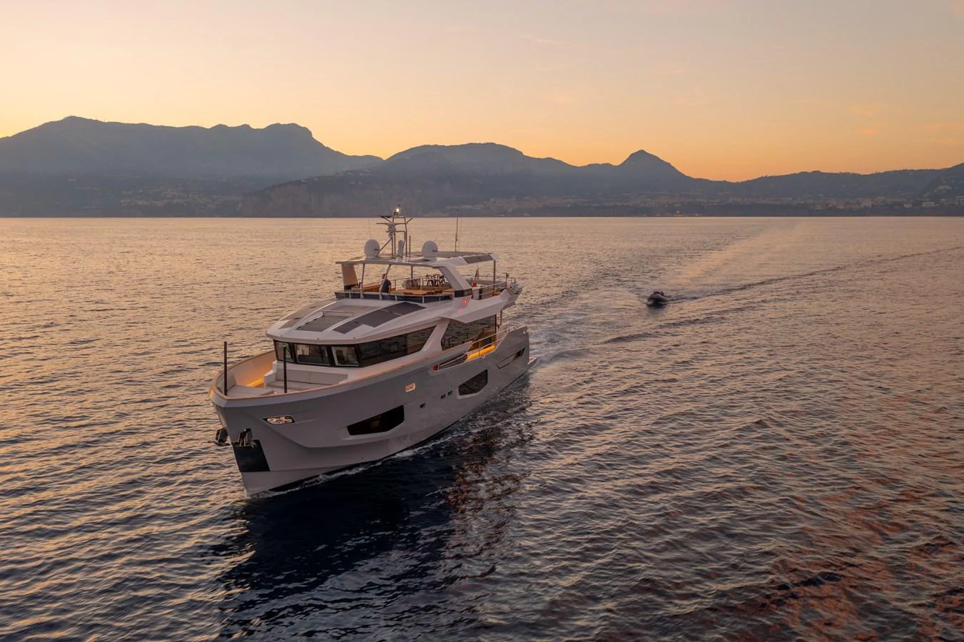 A luxurious yacht cruises gracefully on calm waters at sunset, with mountains in the background. aboard KAMOKA Yacht for Sale