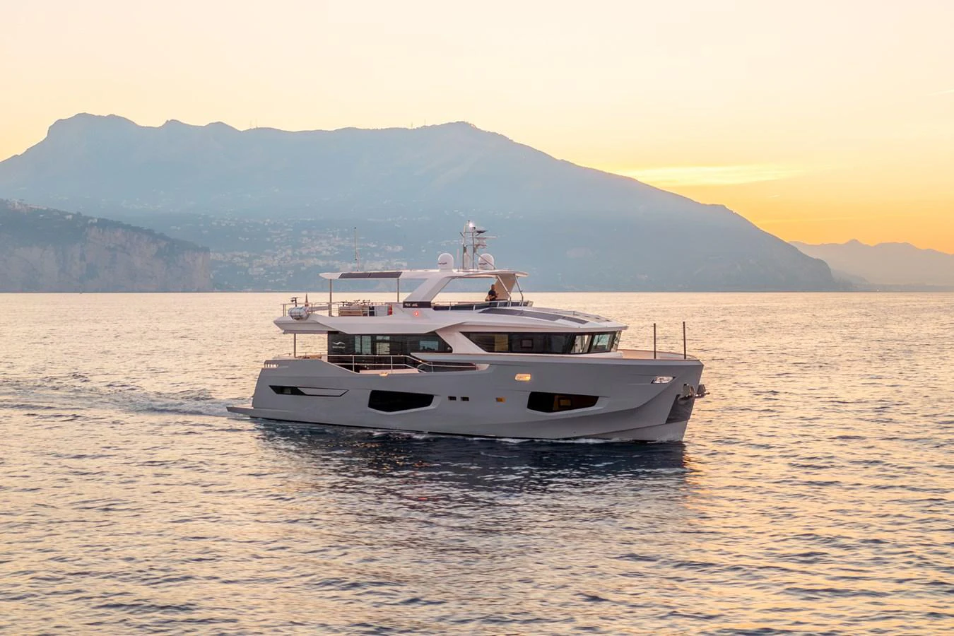 A sleek yacht cruises gracefully on calm waters against a stunning mountainous backdrop at sunset. aboard KAMOKA Yacht for Sale