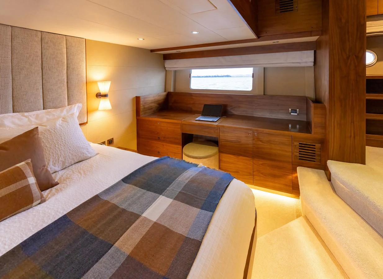 The stateroom features a cozy bed with a plaid blanket, a wooden desk, and a window offering a serene view. aboard DUTCHESS Yacht for Sale
