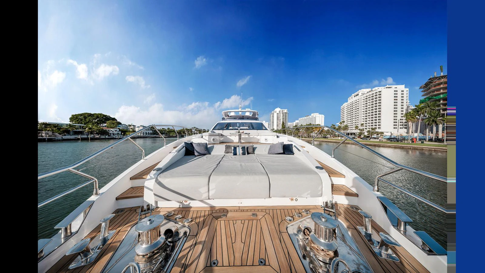 The yacht's foredeck features a spacious sunbathing area with plush cushions, set against a backdrop of a serene waterfront and modern buildings. aboard CLOUD 9 Yacht for Sale