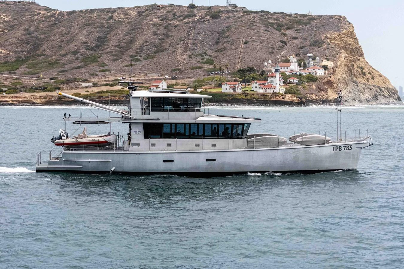 A sleek, modern yacht cruises along a scenic coastline with rugged cliffs and charming houses in the background. aboard UGLY BETTY Yacht for Sale